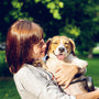 smiling woman standing and carrying short-coated brown and white dog in green field surrounded with tall and green trees
