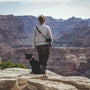 man in gray hoodie and black pants standing on rock with black labrador retriever on top